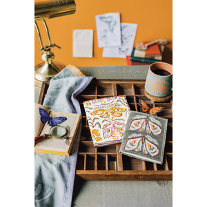 A desk with two Nocturna block print notebooks, an Imprint Mug, and a Jacquard Dishtowel by Danica Studio featuring a moth design, next to a lamp and a magnifying glass.